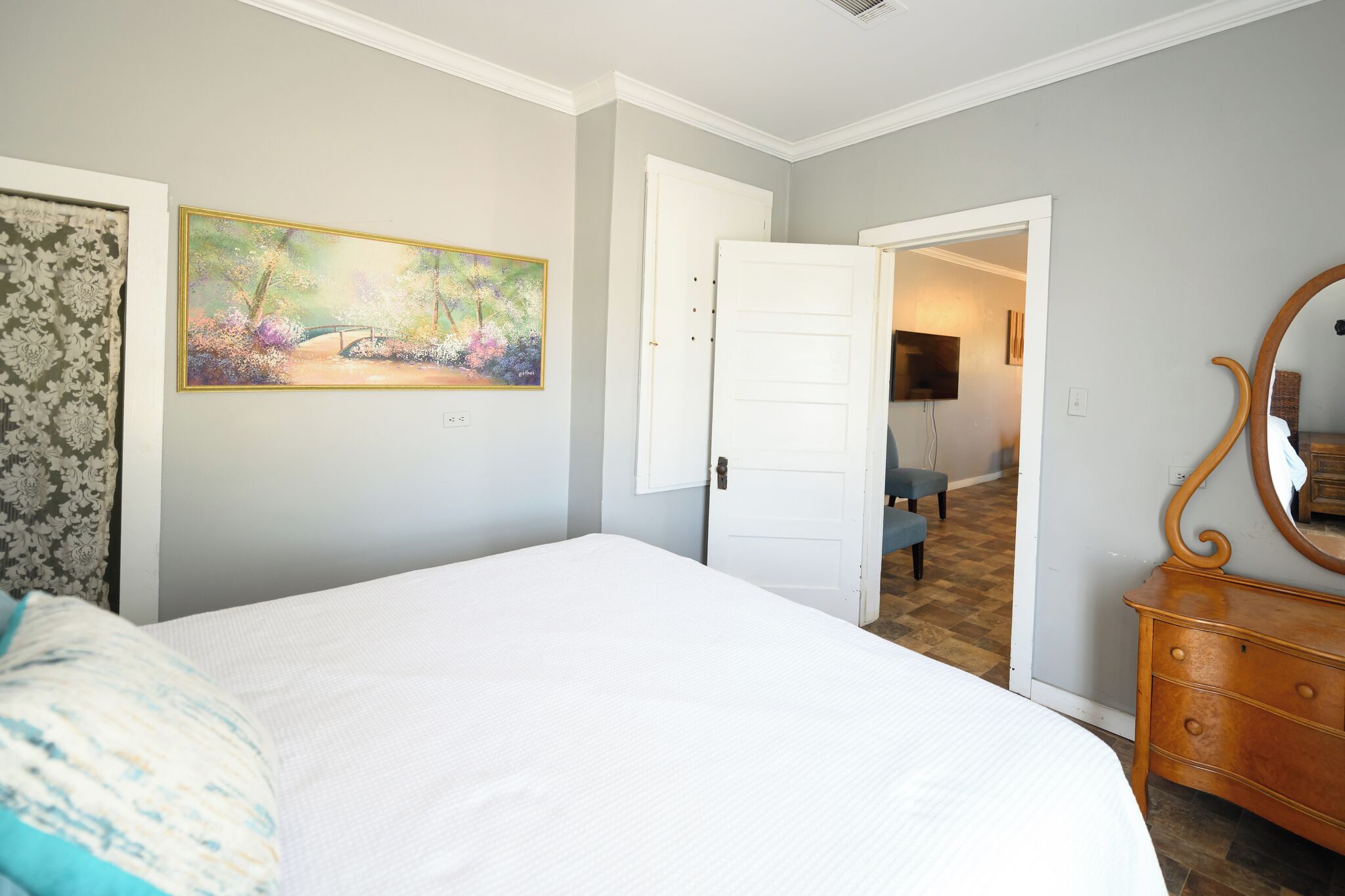 A bedroom with a bed, a dresser, a painting, and a door leading to another room.