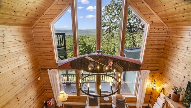 A cozy cabin with wooden walls, a central seating area with two chairs, a fireplace, and large windows offering a scenic view. - Mtn View + Pond w/ fishing + Hot Tub + Firepit + EV charger (Ranger)