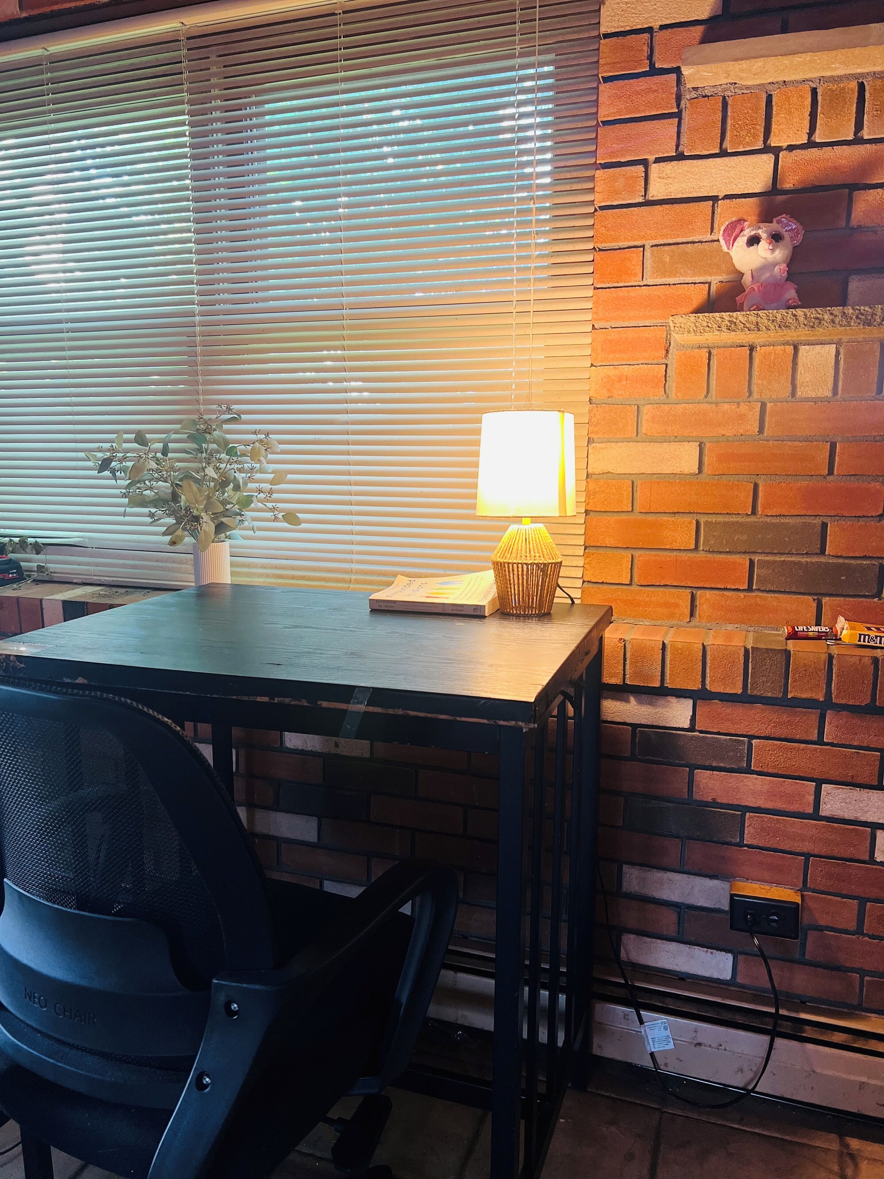 A room with a brick wall, a window with blinds, a table with a lamp, a chair, and a small shelf with a teddy bear.