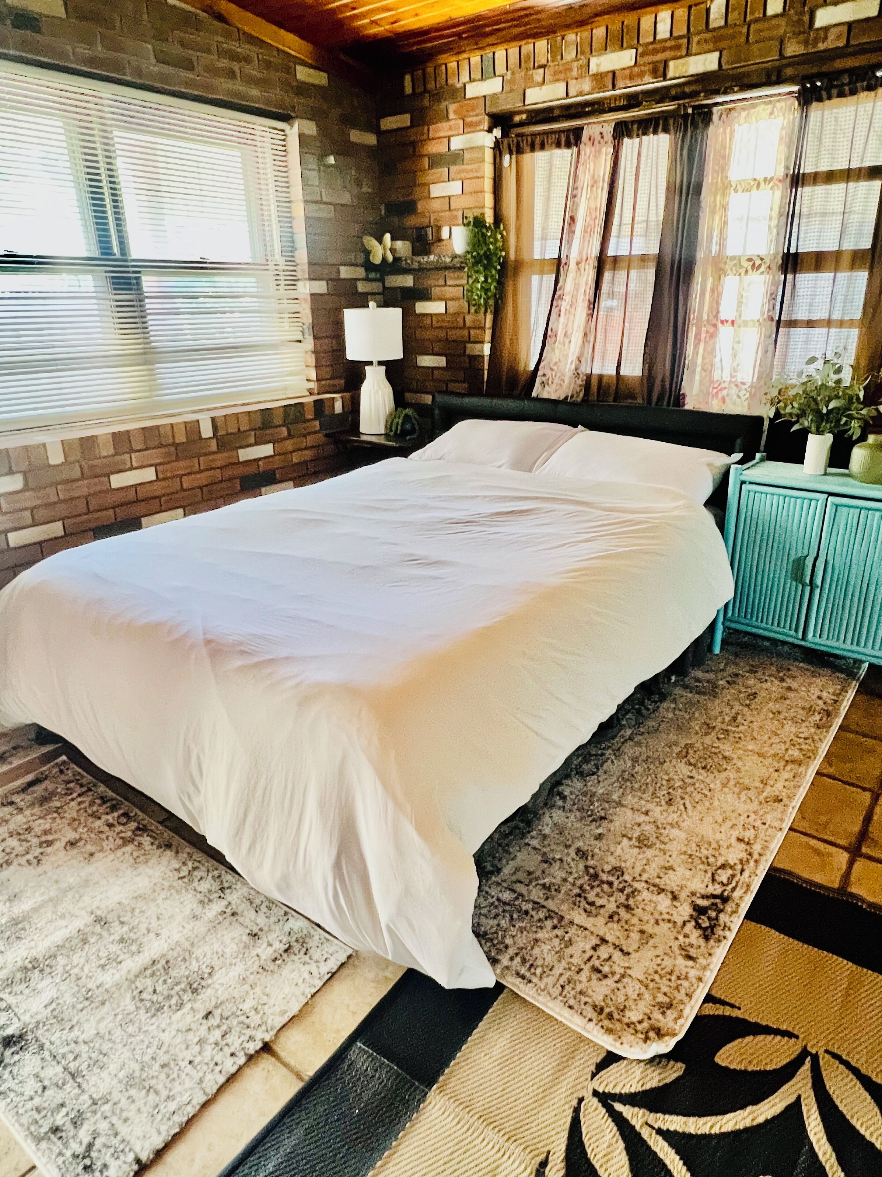 Une chambre avec un grand lit, un mur en briques, une fenêtre avec des stores, une table de chevet avec une lampe et une petite table de nuit avec une plante.