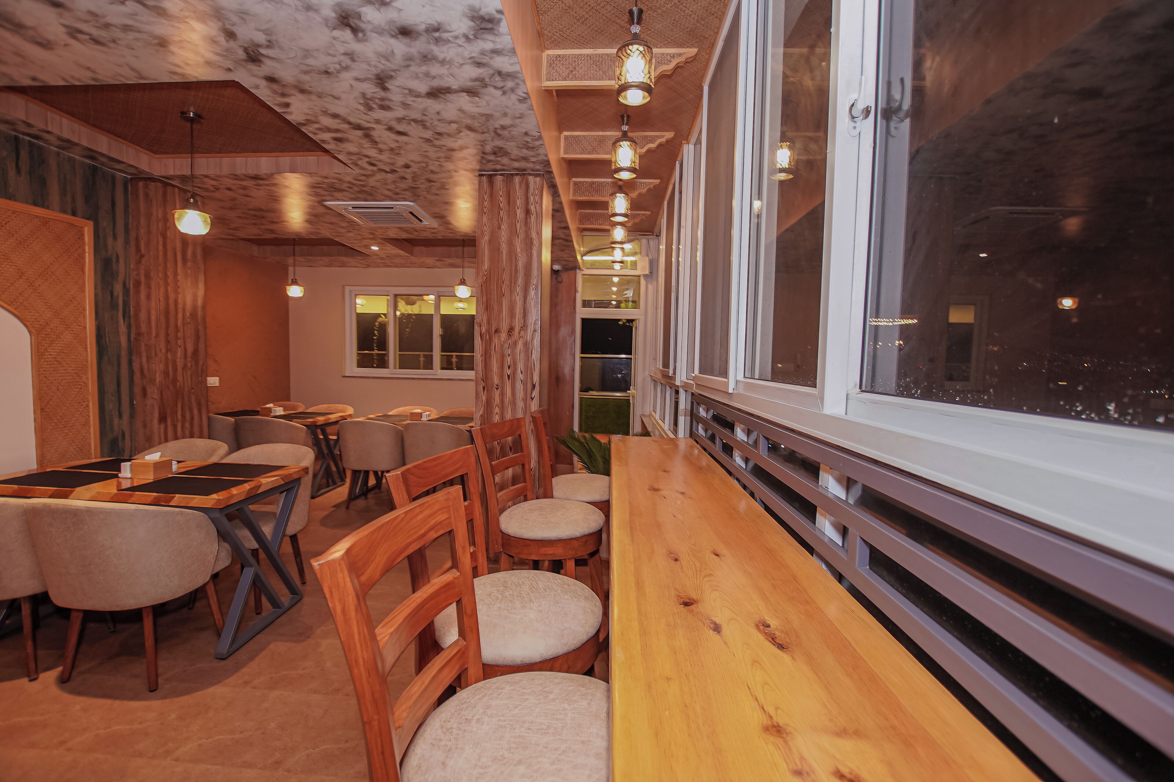 A restaurant interior with wooden tables and chairs, pendant lighting, and large windows.
