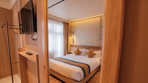 Habitación de hotel con piso de madera, cama con ropa blanca y almohadas marrones, televisor montado y armario de madera.