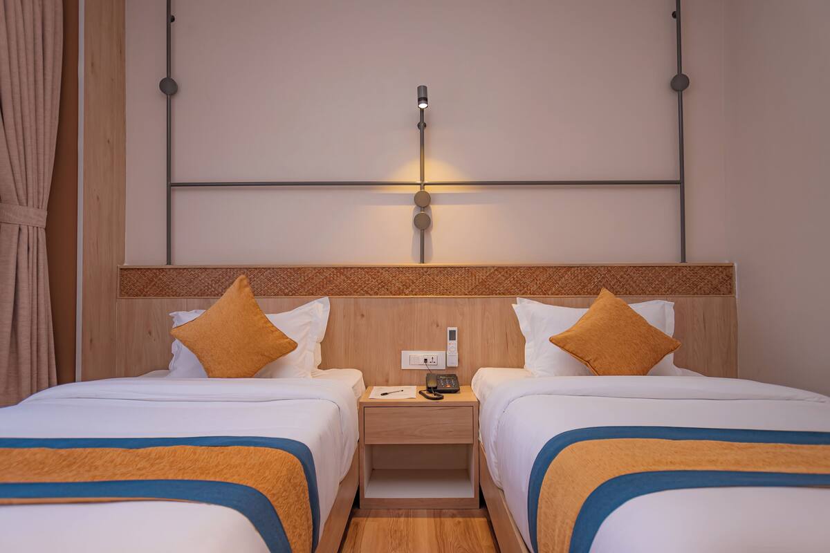 A hotel room with two beds, a wooden headboard, a nightstand with a phone and a lamp, and a wall-mounted light fixture.