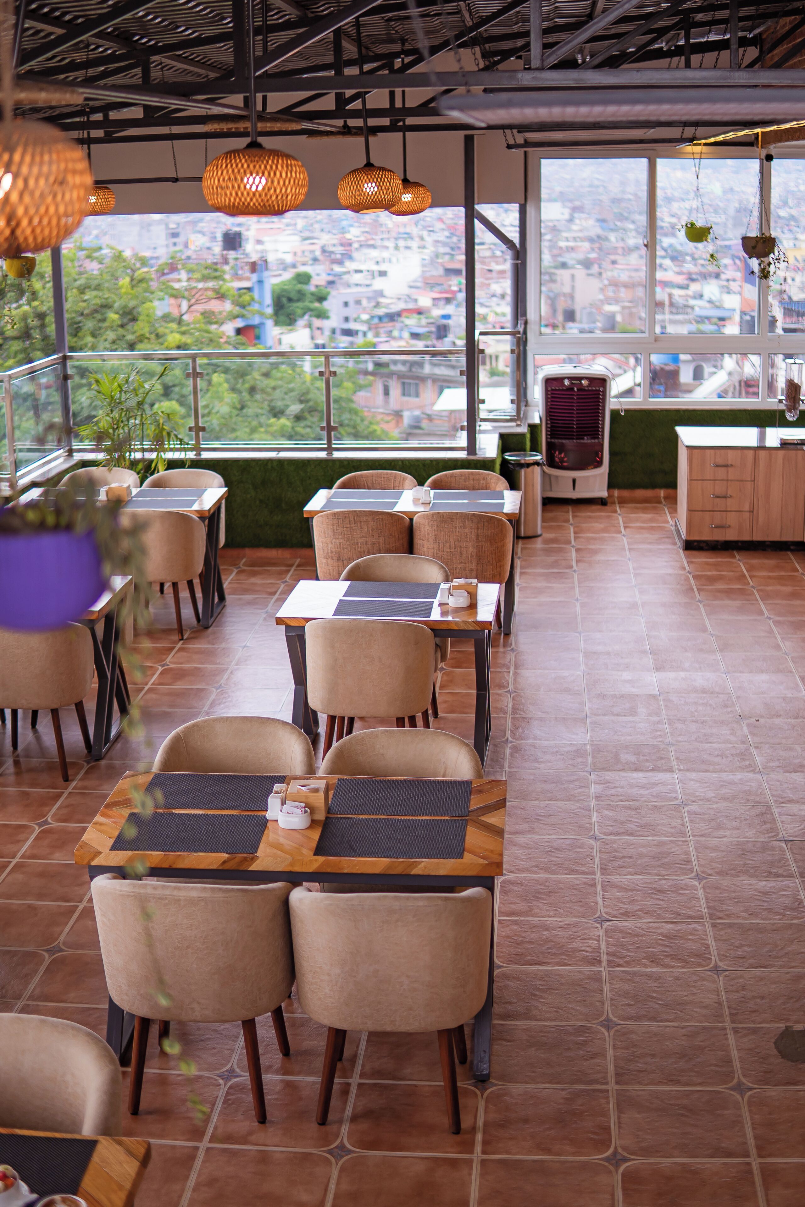 A restaurant with high tables and chairs, large windows offering a city view, hanging pendant lights, and a visible kitchen area.