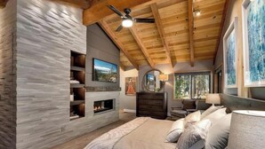 A bedroom with a stone accent wall, a fireplace, a bed with multiple pillows, a dresser, and a ceiling fan. - The Lodge at Wildwood (South Lake Tahoe)