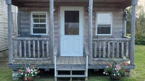Une petite cabane en bois avec un porche à l’avant, deux fenêtres et une porte. Sur le porche, on trouve des plantes en pot, certaines fleuries, et un drapeau américain.