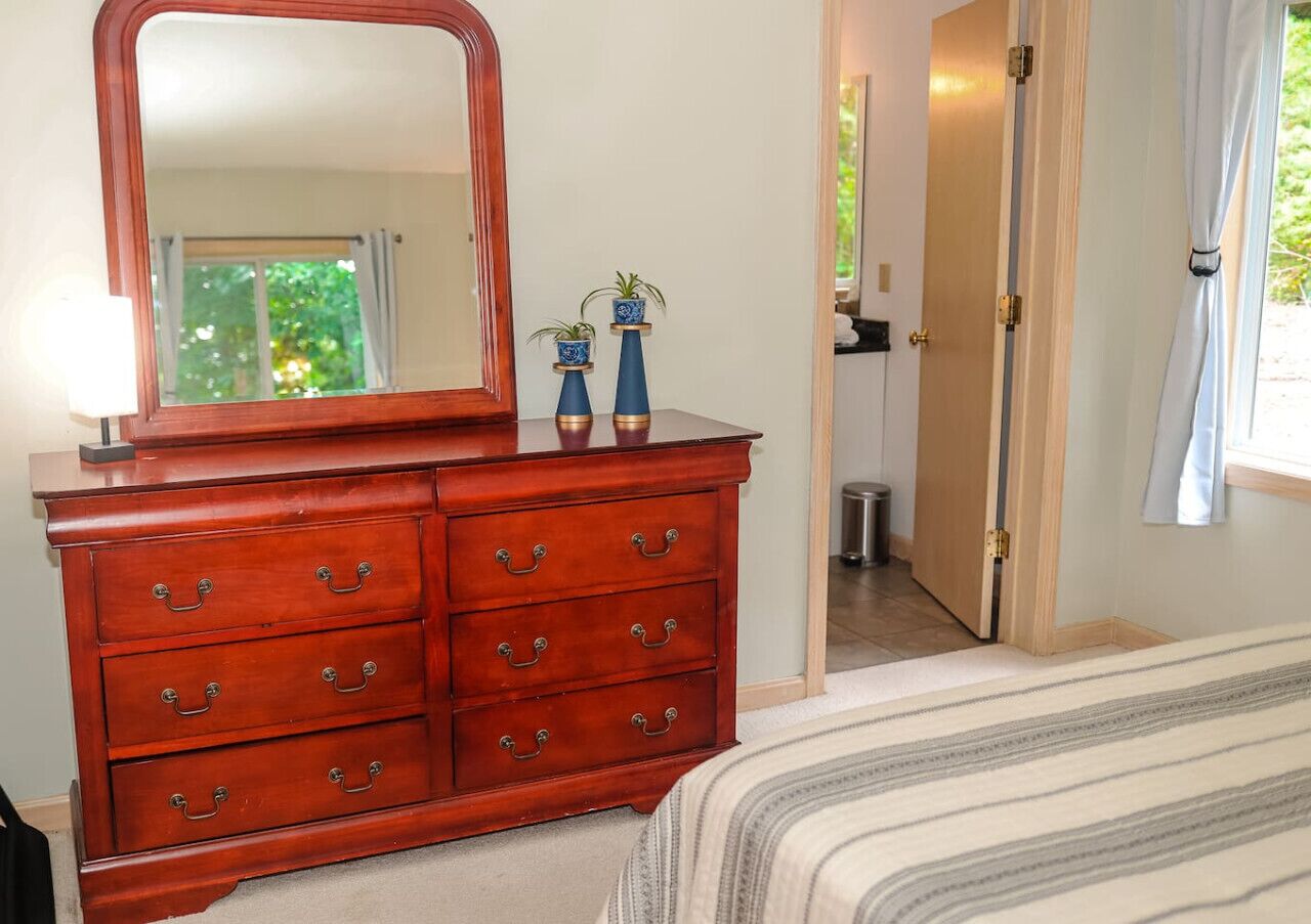 Een slaapkamer met een houten dressoir, een bed met gestreept beddengoed, een spiegel en twee potplanten op het dressoir.