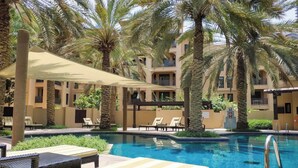 Zona de piscina con sillones de descanso, palmeras y un edificio con balcones.