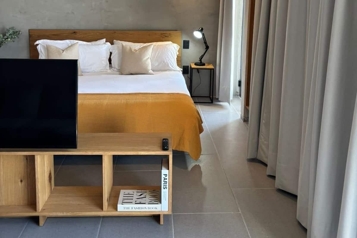 Una habitación de hotel moderna con una cama de madera, un televisor sobre un mueble de madera, una lámpara de noche y un ventanal grande con cortinas.
