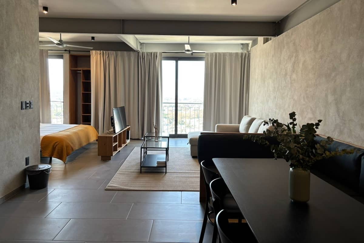 Una habitación de hotel moderna con sofá, mesa de comedor, televisor y balcón con cortinas.