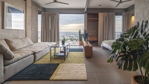 Una sala moderna con un sofá, una mesita de centro y una planta grande. Hay dos ventiladores de techo y una vista de la ciudad a través de ventanas amplias.