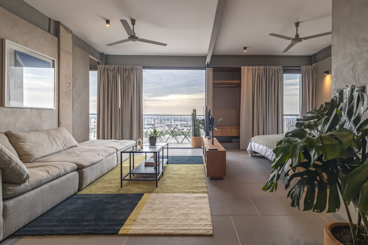 Una sala moderna con un sofá, una mesita de centro y una planta grande. Hay dos ventiladores de techo y una vista de la ciudad a través de ventanas amplias.