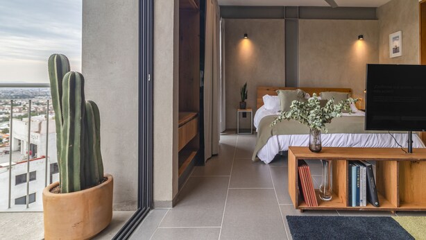 Un dormitorio moderno con cama, mesita de noche y televisor. Se aprecia un ventanal con vista a la ciudad.