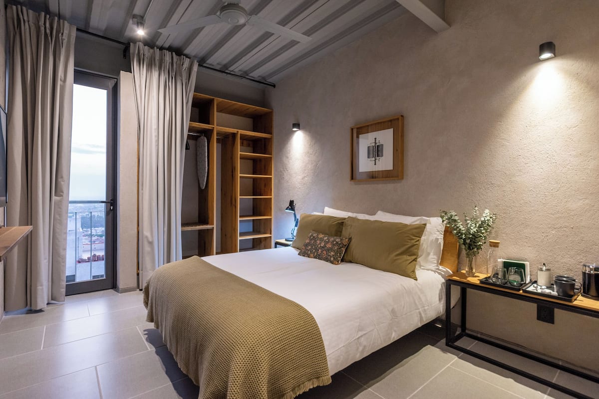 Un dormitorio con una cama grande, un armario de madera, una mesita de noche con un florero y vistas a la ciudad desde un balcón.