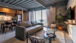 Una sala moderna con sofá, mesa de centro y sillones. Hay una zona de cocina con armarios de madera y un refrigerador. Un ventanal grande proporciona luz natural y vistas de la ciudad.