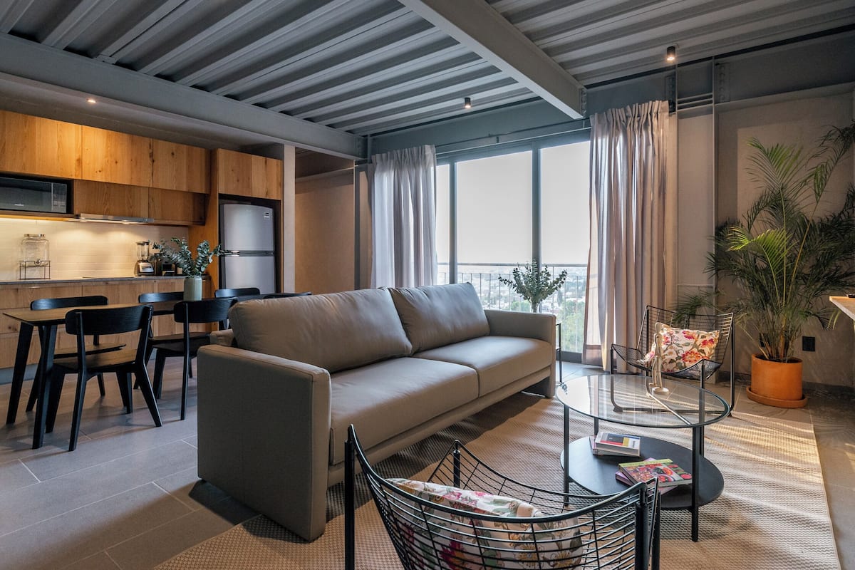 Una sala moderna con sofá, mesa de centro y sillones. Hay una zona de cocina con armarios de madera y un refrigerador. Un ventanal grande proporciona luz natural y vistas de la ciudad.