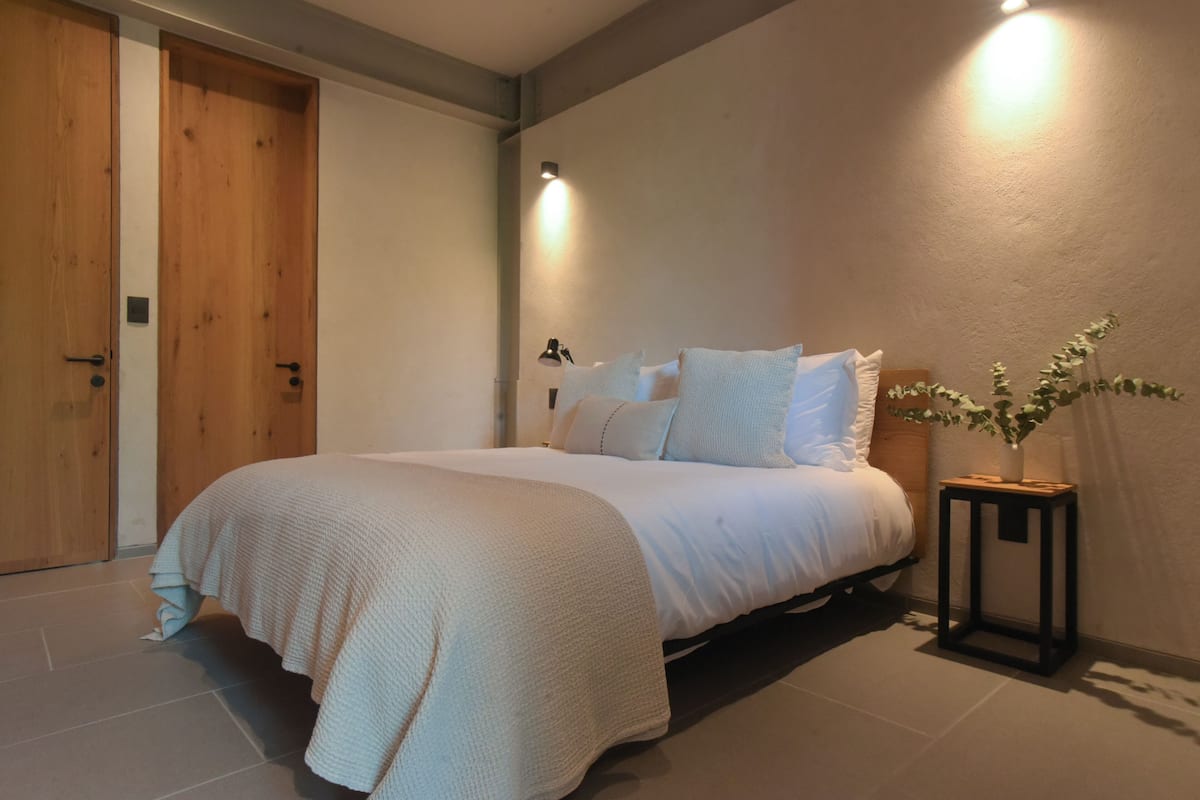 Un dormitorio con una cama, una mesita de noche, una lámpara de pared y una puerta.