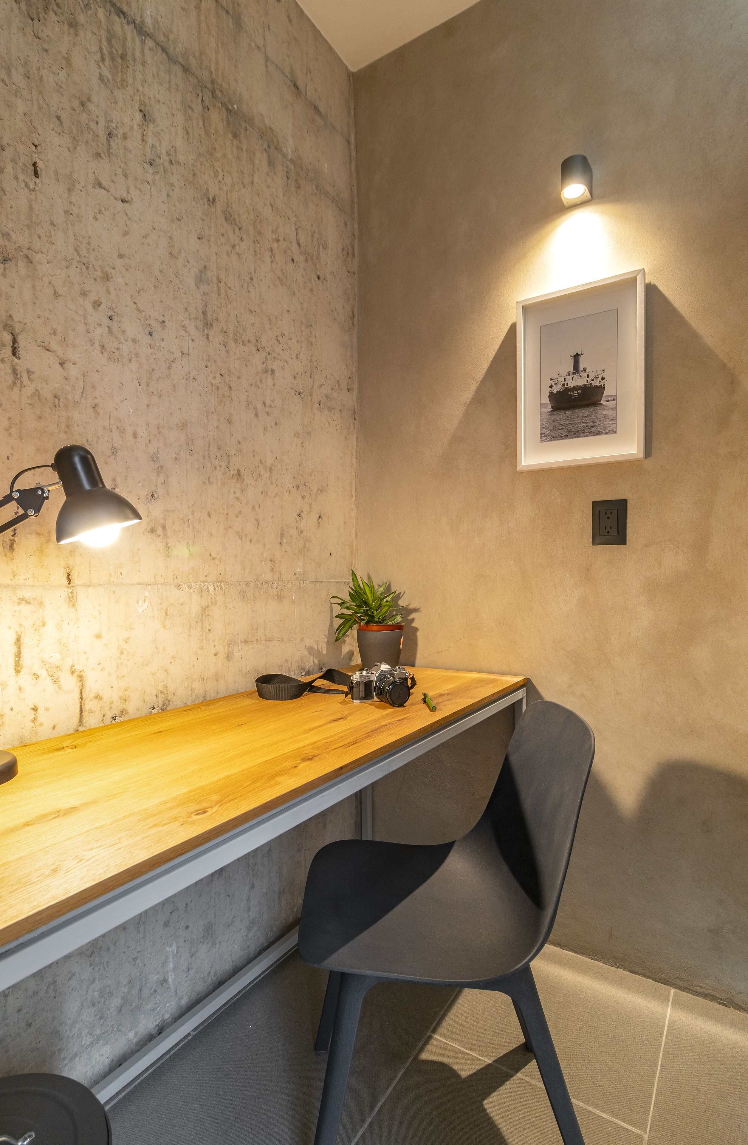 A workspace with a concrete wall, a wooden desk, a black chair, a desk lamp, a framed picture, and a potted plant.