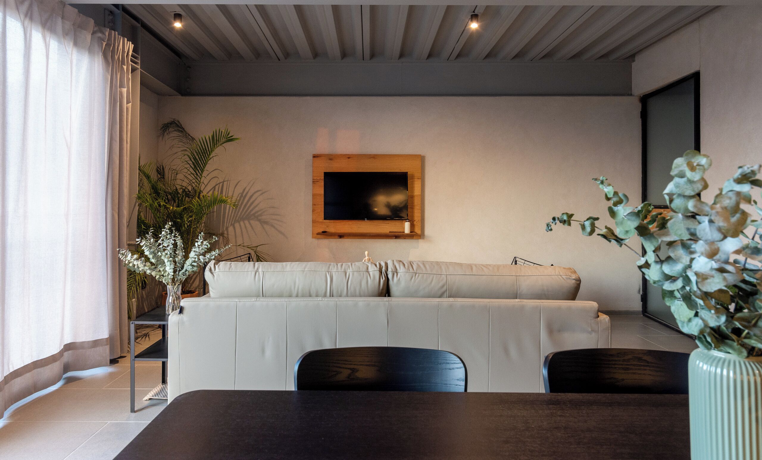 A modern living room with a sofa, a TV mounted on the wall, a dining table, and potted plants.