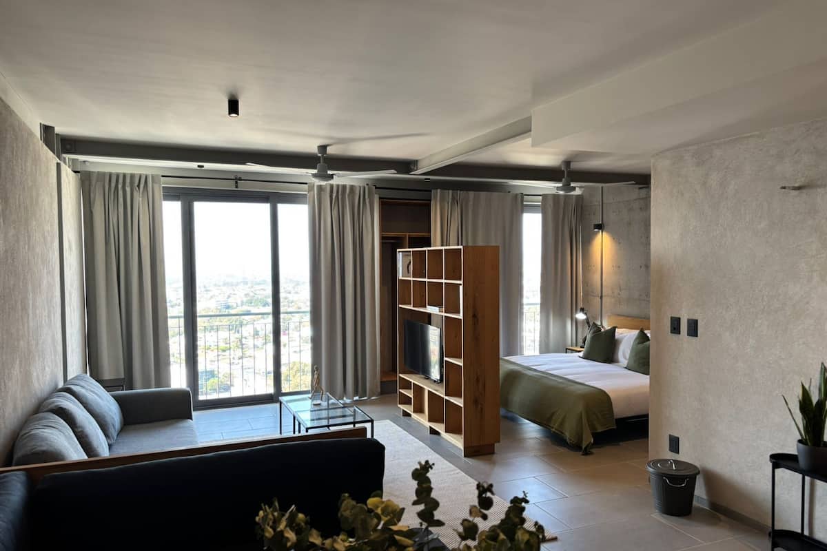 Una habitación de hotel moderna con un ventanal grande, un sofá, una cama, una estantería de madera y una mesita.