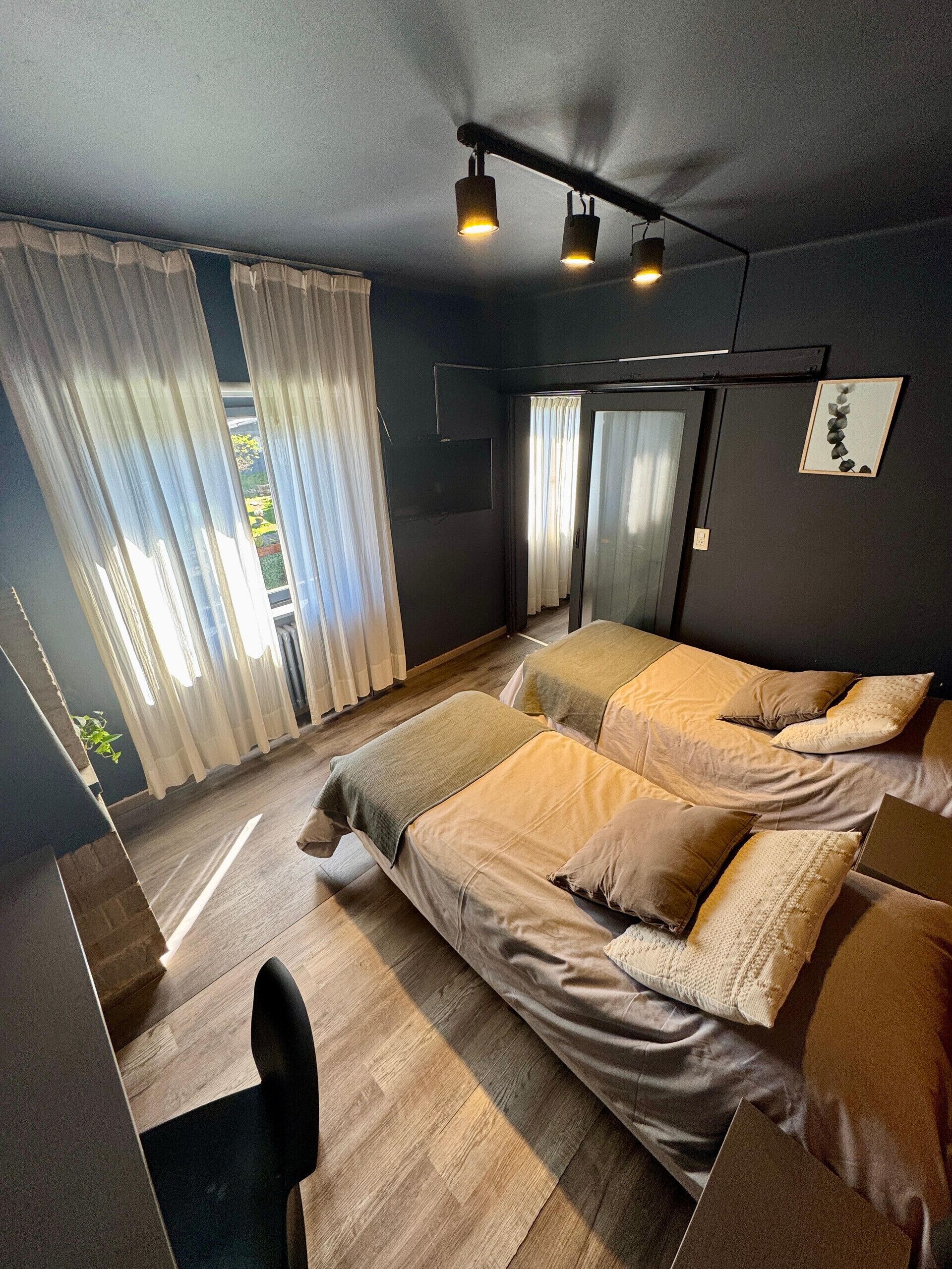 Un dormitorio moderno con una cama grande, un escritorio y una silla. Se ve una ventana con cortinas, una puerta y un cuadro colgado en la pared.