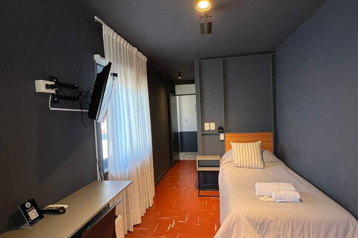 A hotel room with a desk, two beds, a bedside table, a nightstand, and a wall-mounted lamp.