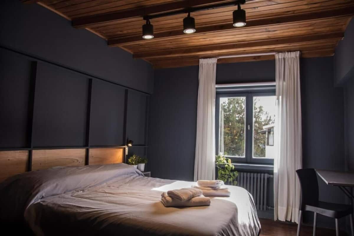 A modern bedroom with a large bed, a desk, and a window with curtains.