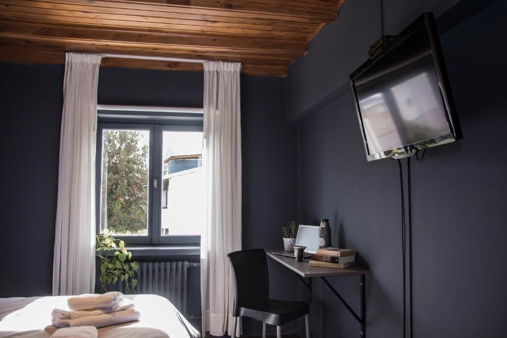 A room with a bed, a desk with a computer, a chair, a TV, and a window with curtains.