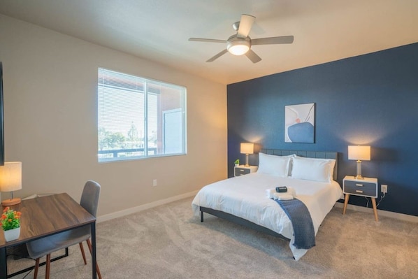 A hotel room with a bed, bedside tables, a desk, a chair, a ceiling fan, and a window.