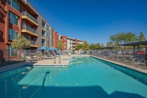 A swimming pool with lounge chairs and umbrellas, surrounded by a fenced area with a multi-story building in the background.