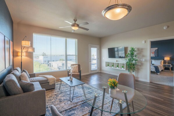 A modern living room with a sofa, coffee table, and chairs, a glass dining table, a ceiling fan, and a TV mounted on the wall. - Prime Spot Apt  | Pool, Gym, Balcony, W/D, Parking (Tempe)