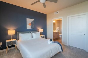 A hotel room with a large bed, two bedside tables with lamps, a ceiling fan, a door to a bathroom, and a small artwork on the wall. - Prime Spot Apt  | Pool, Gym, Balcony, W/D, Parking (Tempe)