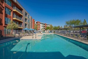 A swimming pool with lounge chairs and umbrellas, surrounded by multi-story buildings with balconies and a clear blue sky. - Prime Spot Apt  | Pool, Gym, Balcony, W/D, Parking (Tempe)