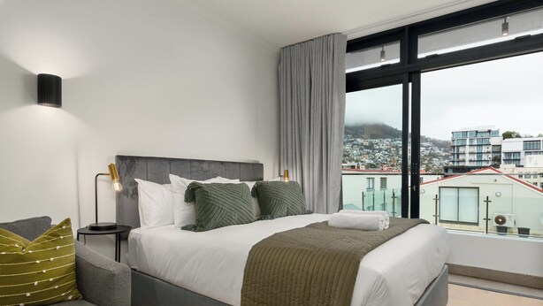A modern bedroom with a large bed, grey and white bedding, a grey sofa, a small side table, and a large window with a city view. - The Sage l Vibrant Sea Point Studio (Cape Town)