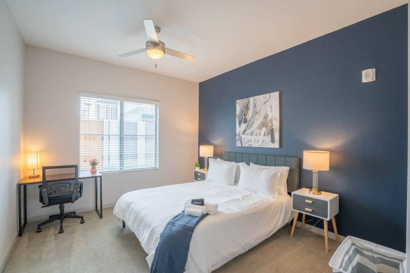 A bedroom with a bed, bedside tables, a desk, a chair, a ceiling fan, and a window.