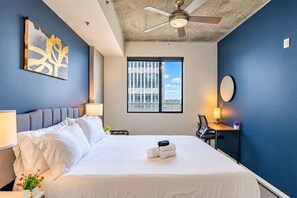 A hotel room with a bed, a ceiling fan, a window with blinds, a desk with a chair, and a wall-mounted mirror. - Arts Downtown PHX | Pool, Gym, Balcony & Parking (Phoenix)