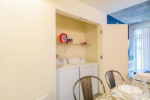 A laundry room with a washing machine and dryer, a shelf with laundry detergent, and a dining area with a table set for two. - Arts Downtown PHX | Pool, Gym, Balcony & Parking (Phoenix)