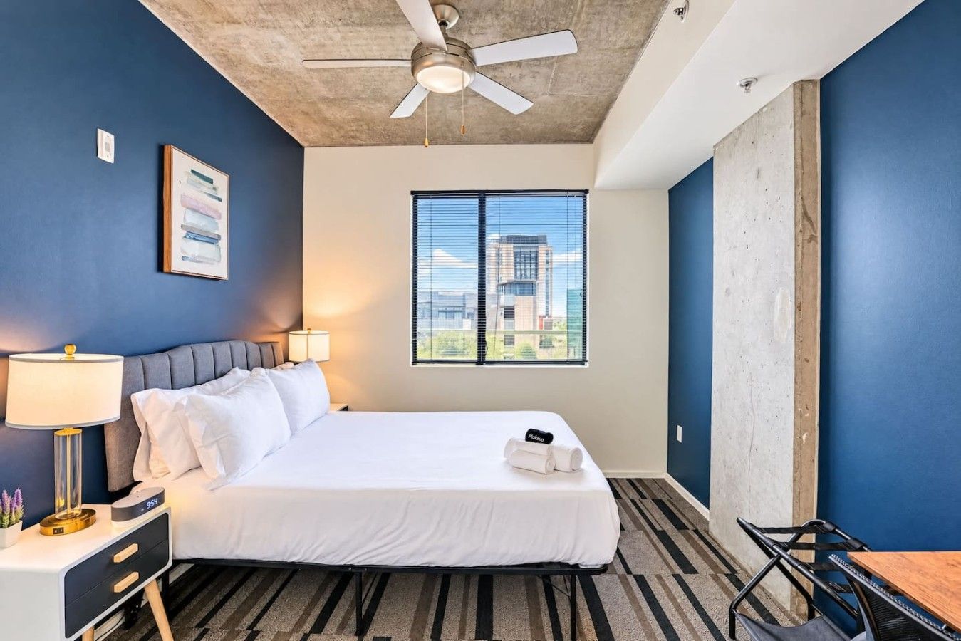 Une chambre d’hôtel comprenant un lit, un bureau, une chaise, un ventilateur de plafond, une fenêtre donnant sur la ville et une lampe.