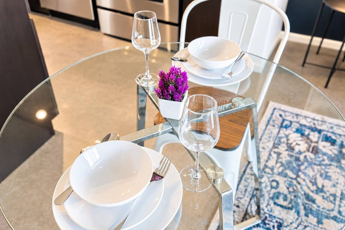 A round glass table set for two with white plates, cutlery, and wine glasses, accompanied by a small purple flower arrangement.