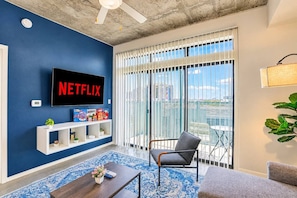 A living room with a large flat-screen TV mounted on a blue wall, a white entertainment unit below it, a grey armchair, a blue patterned rug, and a balcony with a view of the city. - PHX DT Apartment with Balcony + Gym + Pool Parking (Phoenix)