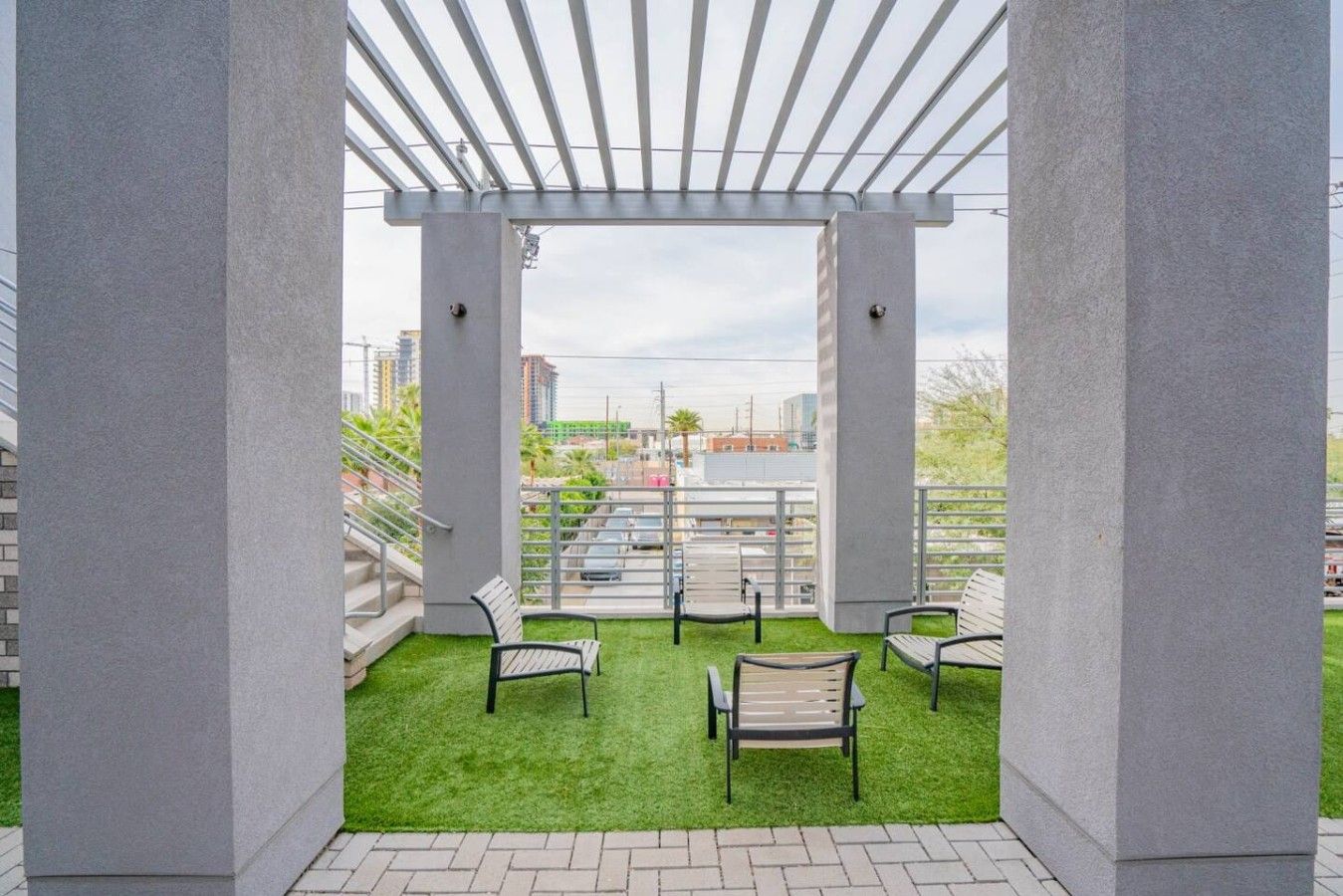 A covered outdoor seating area with artificial grass and metal chairs, overlooking a cityscape.