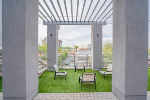 Un espace extérieur aménagé avec une zone de repos couverte, dotée d’un revêtement de sol en gazon synthétique et de chaises en métal, offrant une vue sur le paysage urbain.