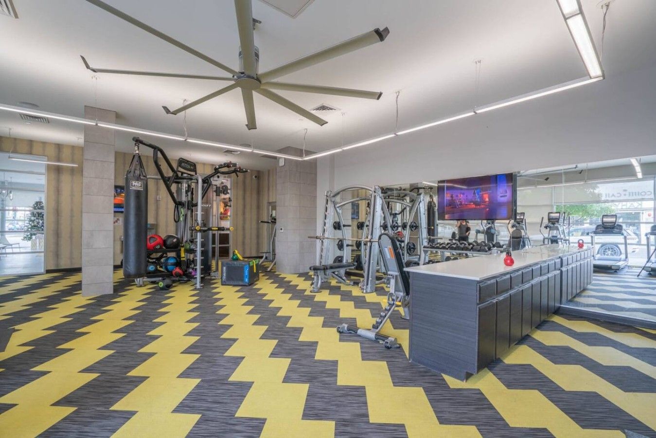 A gym with exercise equipment, a central weight bench, and a large TV screen.