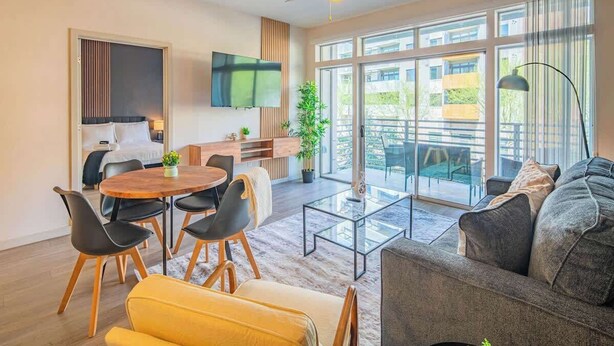 Ein modernes Wohnzimmer mit Sofa, einem Esstisch mit Stühlen, einem an der Wand befestigten Fernseher und großen Fenstern mit Blick ins Freie.