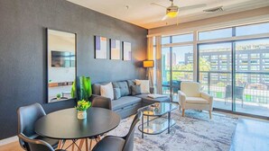 A modern living room with a grey sofa, a round dining table, and a glass coffee table. There is a large window with a view of buildings and greenery outside.