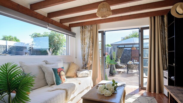 A cozy living room with a sofa, a wooden coffee table with flowers, and a view of the outside patio area. - Boutique Boho Glam Villa With Private Pool (Yaiza)