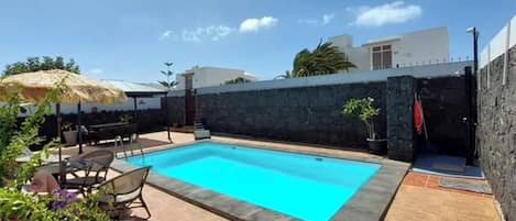 A rectangular swimming pool with a stone border, surrounded by a patio with outdoor furniture and potted plants.