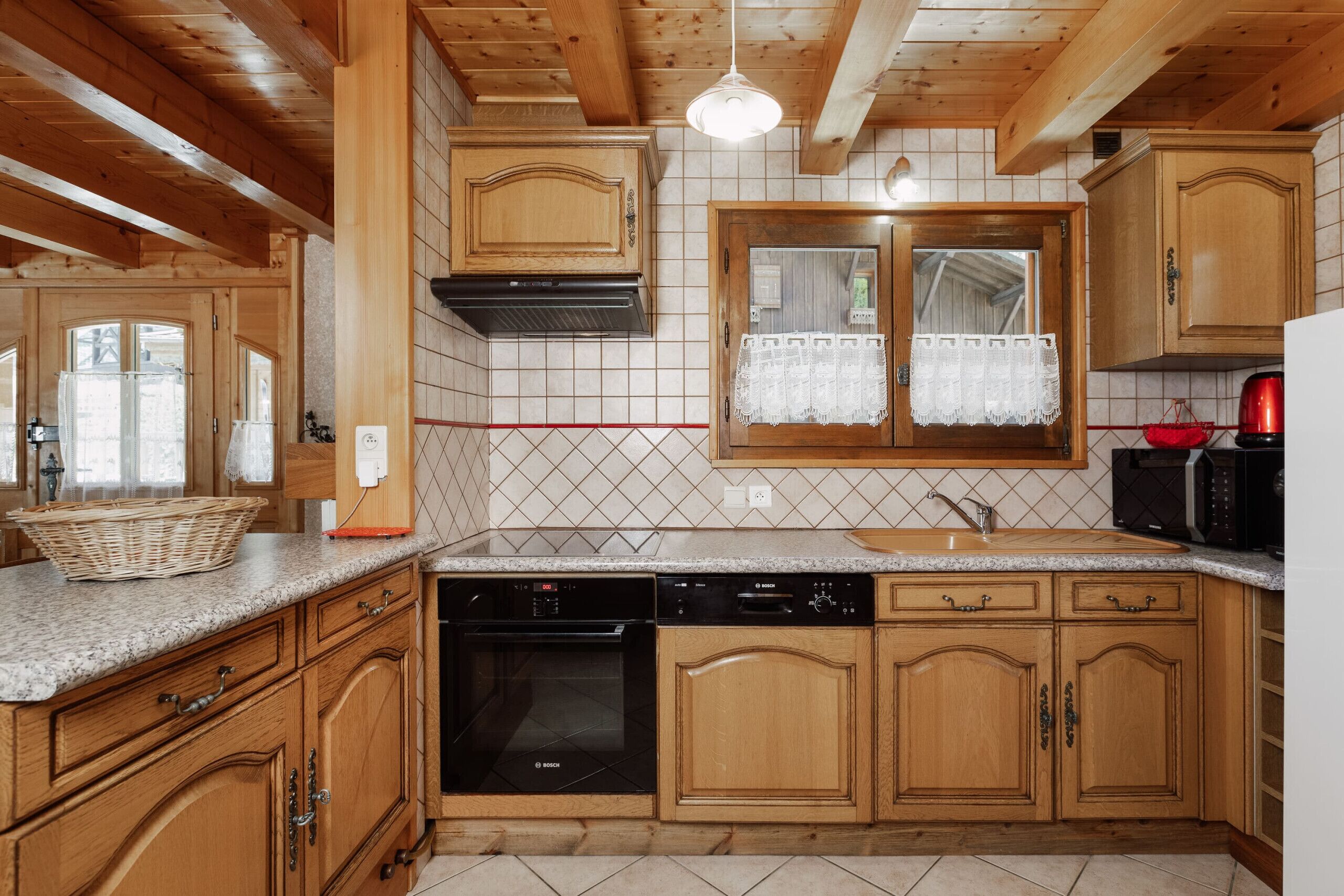 Una cocina con armarios de madera, un lavavajillas Bosch, un fregadero y una ventana con cortina de encaje.