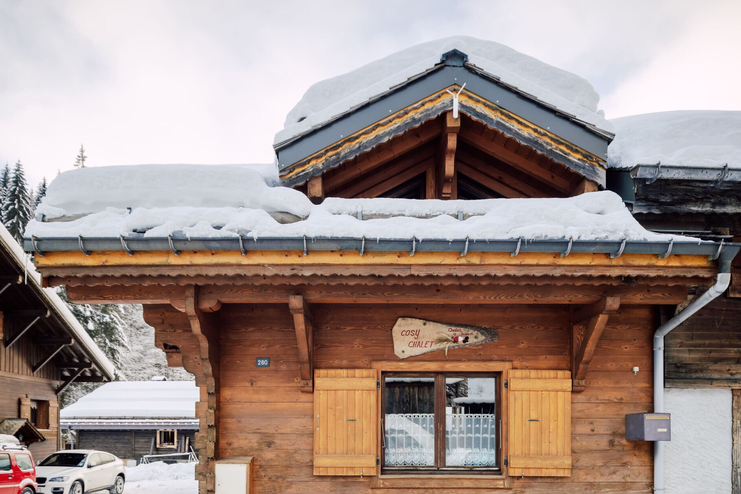Een houten chalet met een sneeuwbedekt dak, een houten deur en een bordje met de tekst 'Cosy Chalet'.