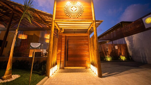 A wooden entrance with a sun-like design, a sign indicating 'Sofit Opening', and a brick pathway leading to the entrance. - Uri Pousada (Maragogi)